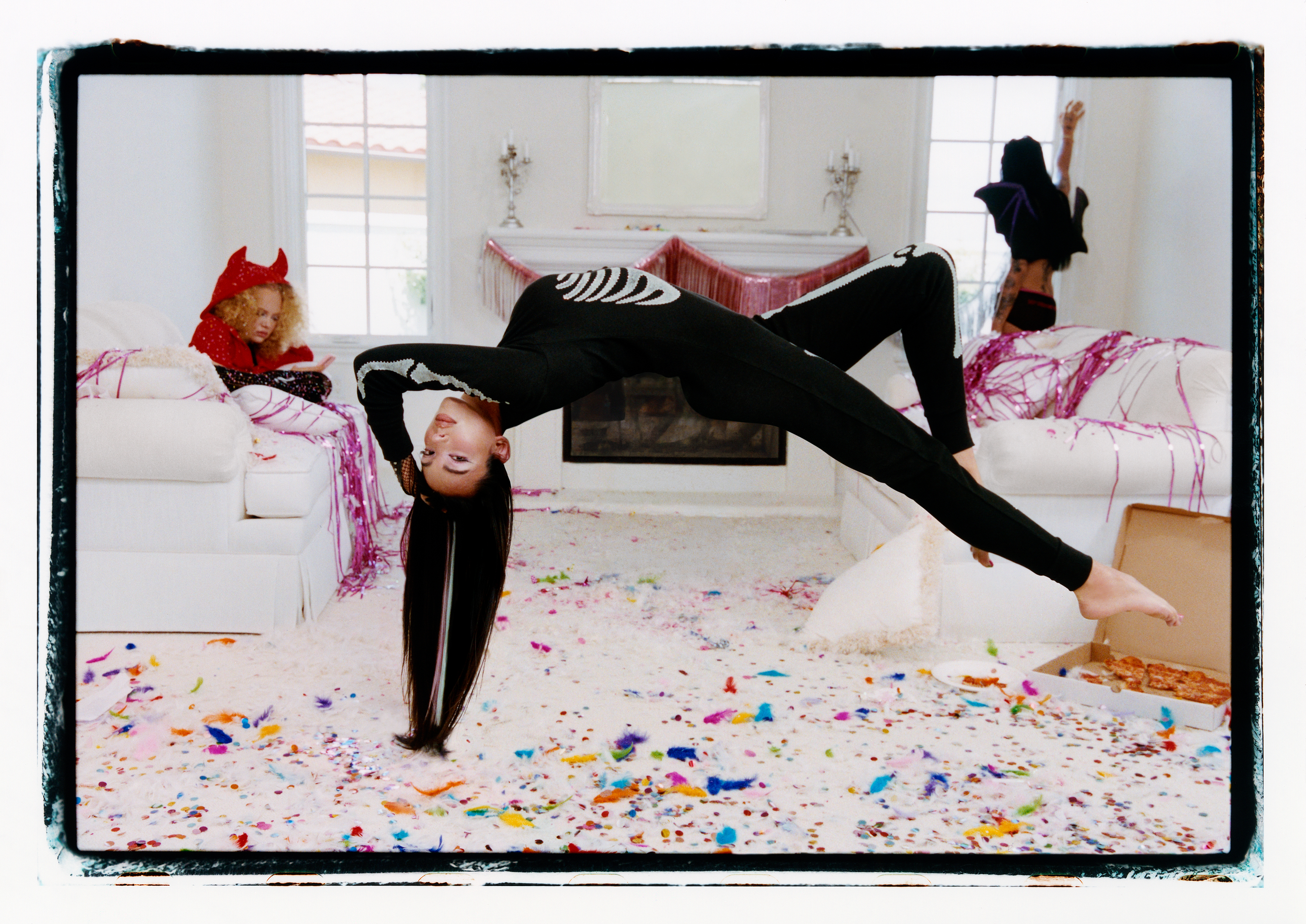 Model in a black one pajama suit levitating over the ground in a white room covered in colorful confetti